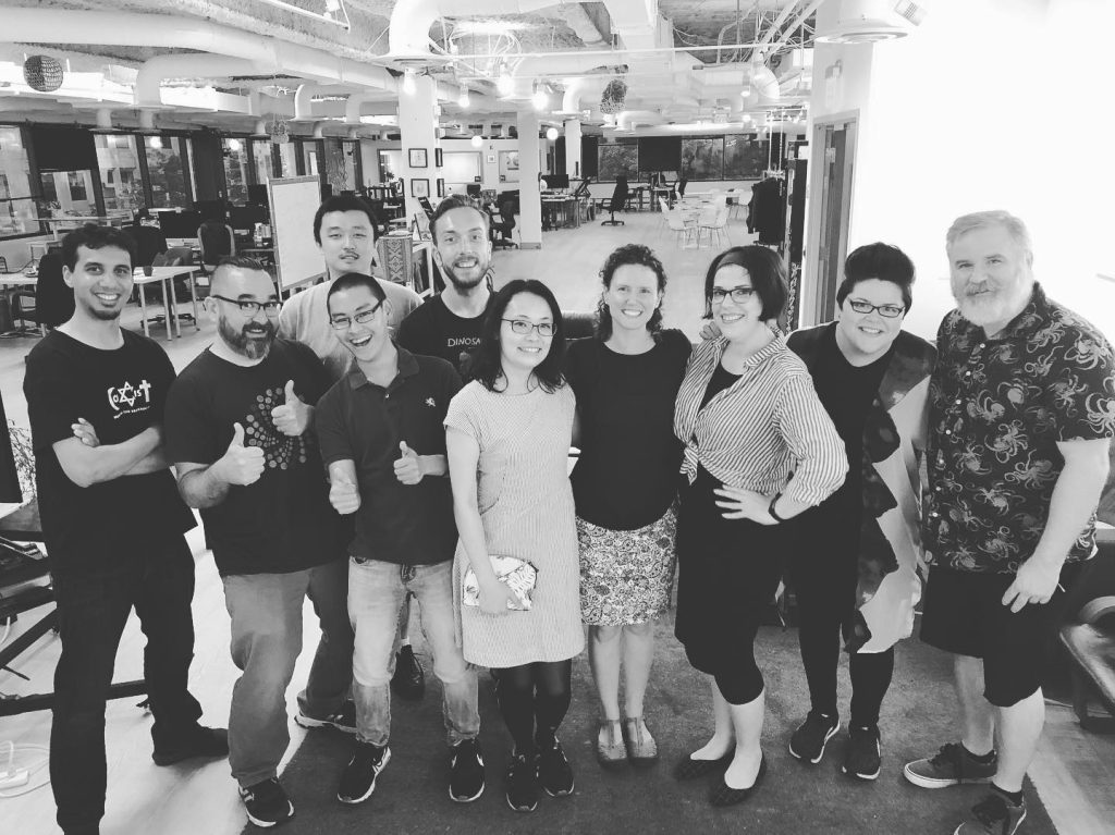A group of friends, standing side-by-side and smiling for the camera. Everyone is happy. Some people have thumbs up. They're standing in a large coworking space (Indy Hall in Philadelphia, PA). SELF Philly was a small business community for freelancers and solo-practitioners.