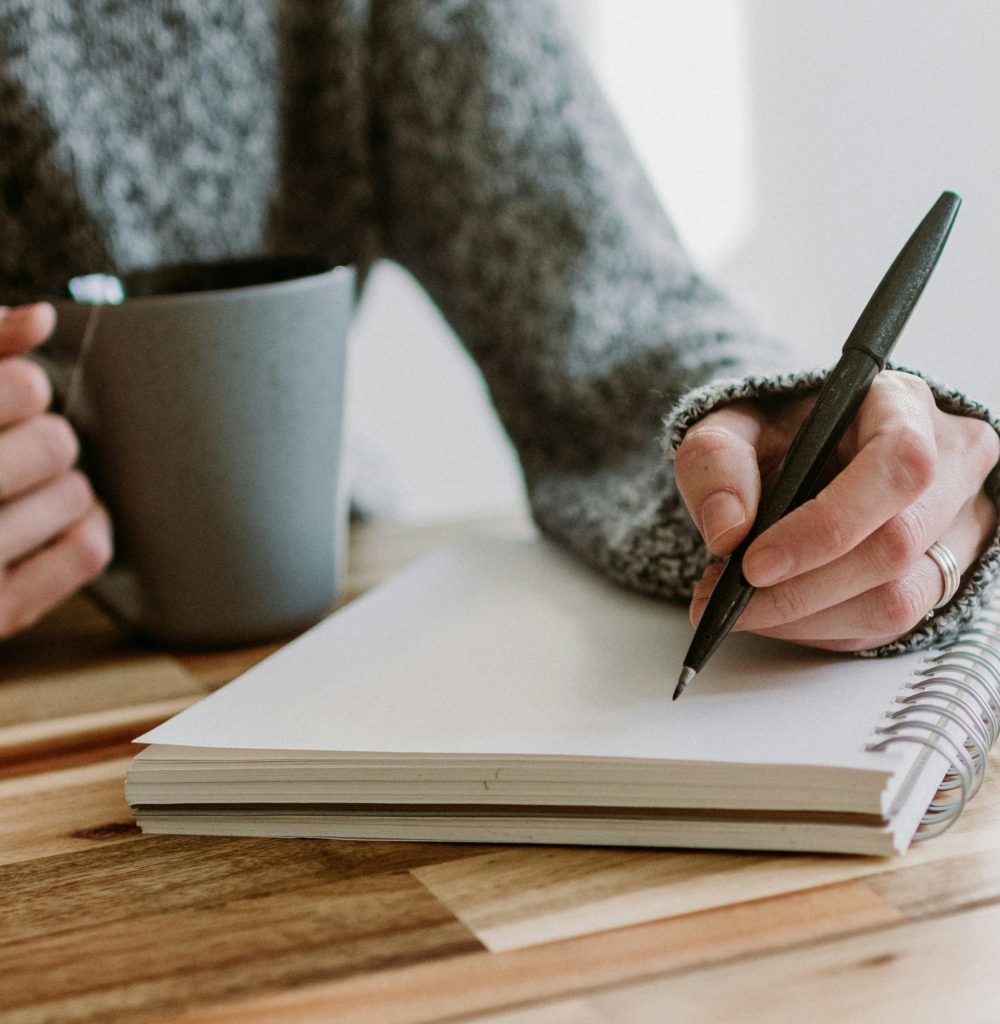 A photo from Unsplash, by Kelly Sikkema. It is of a person writing on a blank notebook with a mug in the other hand. The person is wearing a cozy gray sweater.